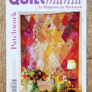 Op de afbeelding: De cover van het tijdschrift Quiltmania toont een kleurrijke patchworkquilt met een verscheidenheid aan patronen en kleuren. De titel van het tijdschrift is paars, met daaronder de woorden "Le Magazine du Patchwork". Een vaas met bloemen, decoratieve potten en een kleine lamp zijn ook zichtbaar.