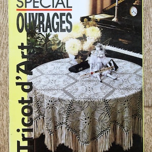 Può includere: Una copertina di rivista con il testo "SPECIAL OUVRAGES" e "Tricot d'Art". La copertina presenta un tavolo rotondo con una tovaglia di pizzo bianco con frange. Un pianoforte, fiori e una bambola sono sullo sfondo. La rivista è sulla maglieria.