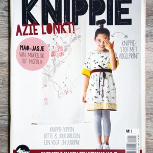 May include: A magazine cover featuring a young girl wearing a white dress with a bird print. The magazine is titled "KnipPie" and the text "Azie Lonkt!" is also visible. The magazine cover features a sewing pattern for a Mao-style jacket.