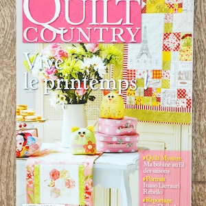 Op de afbeelding: De cover van het tijdschrift "Quilt Country" toont een lentethema. De titel staat in grote witte letters op een roze achtergrond. Een boeket bloemen staat in een witte vaas. De cover bevat ook een quilt met een Eiffeltoren-ontwerp en de tekst "Vive le printemps!"