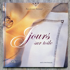 Pode incluir: Uma capa de livro com fundo branco e uma sobreposição de tecido bege. O título do livro é "Jours sur toile" e a autora é Sandrine Sitaud. O livro é sobre bordado e apresenta um logotipo DMC.