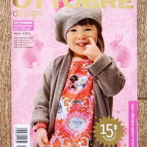 May include: A magazine cover with a child in a grey beret and cardigan. The cover reads "OTTOBRE design" and "OTTOBRE kids fashion 62-170 cm". The background is pink with pink bunny illustrations. The magazine celebrates its 15th anniversary.