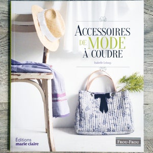 Può includere: Copertina del libro "Accessoires de Mode à Coudre" di Isabelle Leloup. Presenta un cappello di paglia, una borsa tote fantasia con fiocco nero e una sedia con un tessuto drappeggiato. Pubblicato da Éditions Marie Claire.