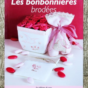Pode incluir: Um livro intitulado "Les bonbonnières brodées" com um fundo rosa e uma caixa branca cheia de rosas cor-de-rosa. A caixa está ao lado de uma bolsa de tecido rosa em forma de coração com uma fita rosa. O livro está sobre uma superfície de madeira.
