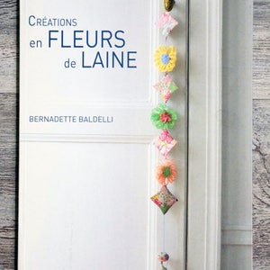 May include: A book titled "Créations en FLEURS de LAINE" by Bernadette Baldelli. The cover features a white background with a blue title and a colourful floral garland hanging from a door.