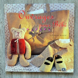 May include: A book cover with the title "Ouvrages pour Bébé" by Sandrine Sitaud. The cover features a brown tote bag, a teddy bear wearing a red vest, a baby dress, and a pair of baby shoes.