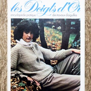 Peut inclure: Une couverture de livre vintage avec le titre "les Doigts d'Or" en lettres bleues. Le livre est une encyclopédie de travaux d'aiguilles et présente une femme portant un pull tricoté dans un décor de forêt.