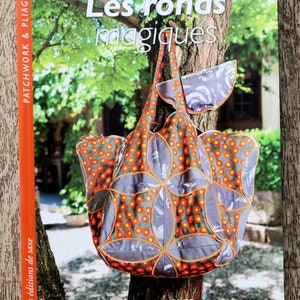 Puede incluir: Un libro titulado "Les ronds magiques" de Bernadette FAVRE. La portada del libro presenta una bolsa de mano de patchwork gris y naranja colgada de una rama de árbol. La bolsa tiene un patrón floral y está hecha con piezas circulares de tela.