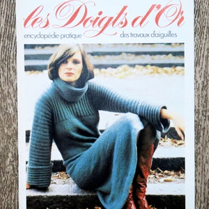 Puede incluir: Una portada de revista con una mujer que lleva un vestido de punto azul y botas rojas. La revista se titula "les Doigts d'Or" y se describe como una enciclopedia de trabajos de aguja.