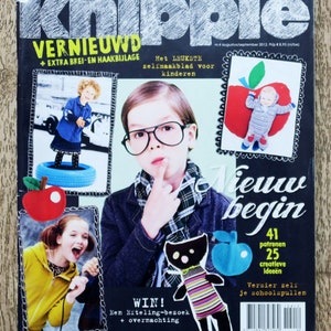 May include: A magazine cover for a Dutch craft magazine called "Knippie" featuring the text "Vernieuwd" and "Het Leukste zelimaakblad voor kinderen". The cover features a child wearing glasses and a plaid shirt, a child wearing a blue shirt and a red apple, and a child wearing a yellow shirt and holding a microphone. The magazine cover also features a black cat with a striped shirt and a red apple.