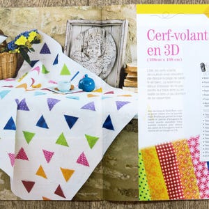May include: A white quilt with colorful triangular patches. The quilt is laid out on a wooden surface. The text "Cerf-volant en 3D (110cm x 108 cm)" is visible in the image.