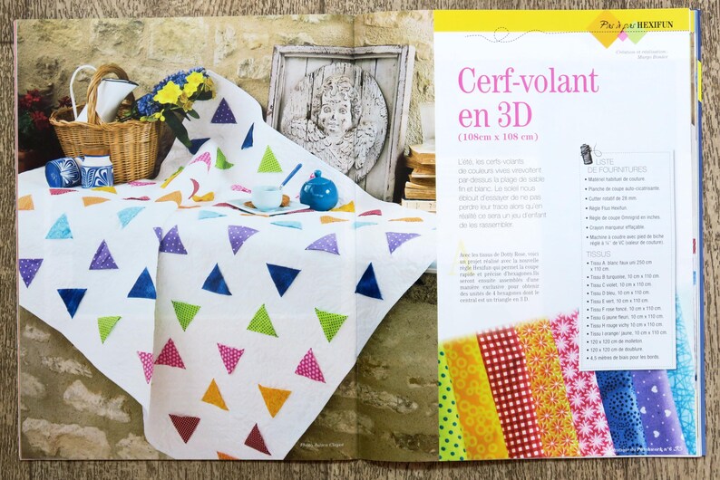 May include: A white quilt with colorful triangular patches. The quilt is laid out on a wooden surface. The text "Cerf-volant en 3D (110cm x 108 cm)" is visible in the image.