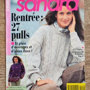Op de afbeelding: Een tijdschriftomslag met een vrouw in een lichtgrijze kabelgebreide trui. De tijdschrifttitel "sandra" is roze. Tekst bevat "Rentrée: 27 pulls" en "Et plein d'ouvrages et d'idées déco !"