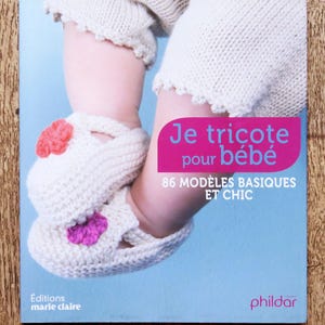 Könnte beinhalten: Buchcover mit dem Titel "Je tricote pour bébé" in Rosa und Weiß. Es zeigt die Beine und Füße eines Babys, das weiße gestrickte Babyschuhe mit rosa und lila Blumenakzenten trägt. Der Hintergrund ist hellblau.