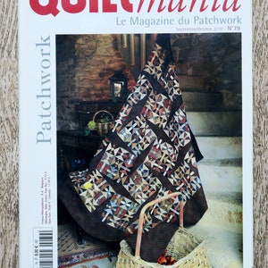 Op de afbeelding: De cover van het tijdschrift "Quiltmania" toont een patchworkquilt met een geometrisch patroon in bruine, beige en blauwe tinten. De titel van het tijdschrift is rood, met "Le Magazine du Patchwork" eronder. Een gevlochten mand staat op de voorgrond.