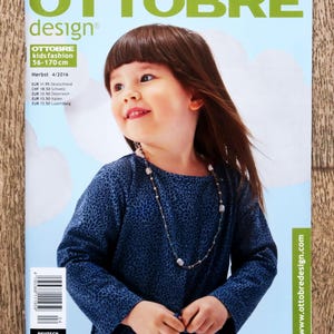 May include: A magazine cover with a child in a blue patterned top and a necklace. The magazine is "OTTOBRE design" with "kids fashion 56-170 cm". The cover also has text in German. The background is light blue.