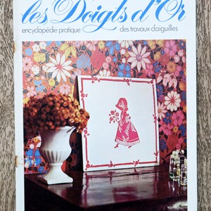 Peut inclure: Une couverture de magazine avec le titre "les Doigts d'Or" et le texte "encyclopédie pratique des travaux d'aiguilles". La couverture présente un fond blanc avec un motif floral rouge, rose et bleu. Un vase en céramique blanc avec un bouquet de fleurs séchées repose sur une table en bois foncé. Un cadre blanc avec une bordure rouge contient une silhouette rouge d'une fille tenant des fleurs.