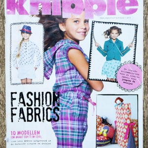 May include: A magazine cover featuring a young girl wearing a plaid shirt and the words "Fashion Fabrics" in bold black letters. The magazine is titled "Knippie" and features 10 sewing patterns for children's clothing.