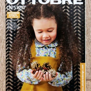 May include: The Ottobre Design magazine cover shows a young girl with brown curly hair holding pine cones. She wears a mustard yellow pinafore over a floral blouse. The magazine title is in large white letters.