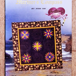 Peut inclure: Couverture de livre "Mariner's Compass" présentant un motif de courtepointe avec des motifs d'éclats d'étoiles jaunes, bleus, roses et violets sur un fond sombre. La couverture comprend le texte "as seen on Quilting from the Heartland" et est présentée par Sharlene Jorgenson.