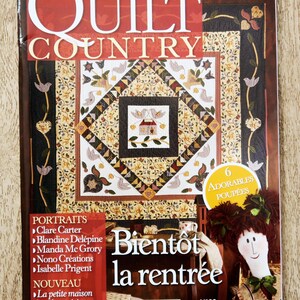 Peut inclure: La couverture du magazine "QUILT COUNTRY" présente un motif de patchwork coloré avec des oiseaux et des motifs floraux. Du texte en français, une poupée et une liste de noms sont également visibles. Le magazine est posé sur une surface en bois.