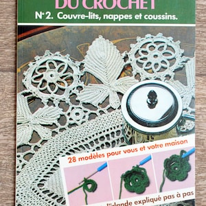 May include: A book titled "L'ÉCOLE DU CROCHET" with the number 2 on the cover. The book features a crocheted doily with a silver lidded container on top. The book cover also includes the text "Couvre-lits, nappes et coussins" and "28 modèles pour vous et votre maison". The book cover also includes the text "le point d'irlande expliqué pas à pas".