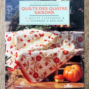 Peut inclure: Couverture de livre "QUILTS DES QUATRE SAISONS" de Linda Seward. Elle présente une courtepointe avec des motifs floraux rouges sur fond blanc. Une grosse citrouille orange et une plus petite sont au premier plan.