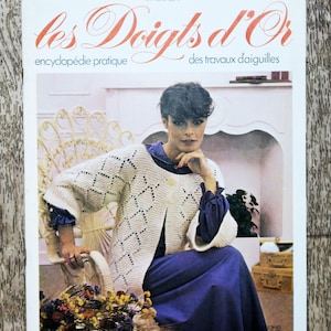 Könnte beinhalten: Ein Magazincover mit einer Frau, die einen weißen Häkelcardigan mit Rautenmuster trägt. Das Magazin heißt "les Doigts d'Or" und wird als eine Enzyklopädie der Nadelarbeit beschrieben.