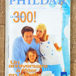 Puede incluir: Una portada de revista que muestra a una familia celebrando un cumpleaños con un pastel azul y blanco. La revista se titula "PHILDAR Creations" y la portada dice "Le numéro 300! Un anniversaire à fêter en famille".