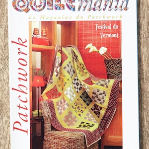 Può includere: La copertina della rivista "Quiltmania" presenta una colorata trapunta patchwork drappeggiata su una sedia di vimini. Il titolo della rivista è in un carattere vibrante e multicolore. La copertina include anche testo in francese e dettagli.