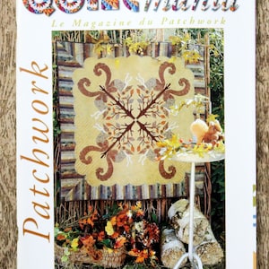 Op de afbeelding: Quiltmania magazine cover met een patchwork quilt met een herfstthema. De quilt is een bruin, oranje en geel ontwerp met een boommotief. De cover toont ook een eekhoorn en een kleine tafel met een wit tafelkleed.