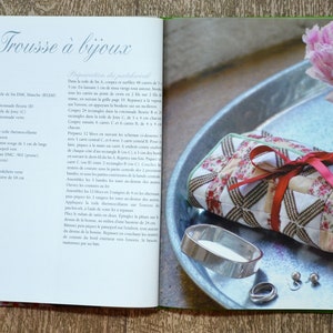 May include: A red and white quilted fabric jewellery roll with a red ribbon tied around it. The roll is sitting on a silver tray with a silver ring and two earrings. The tray is on a wooden surface with a pink flower in the background.