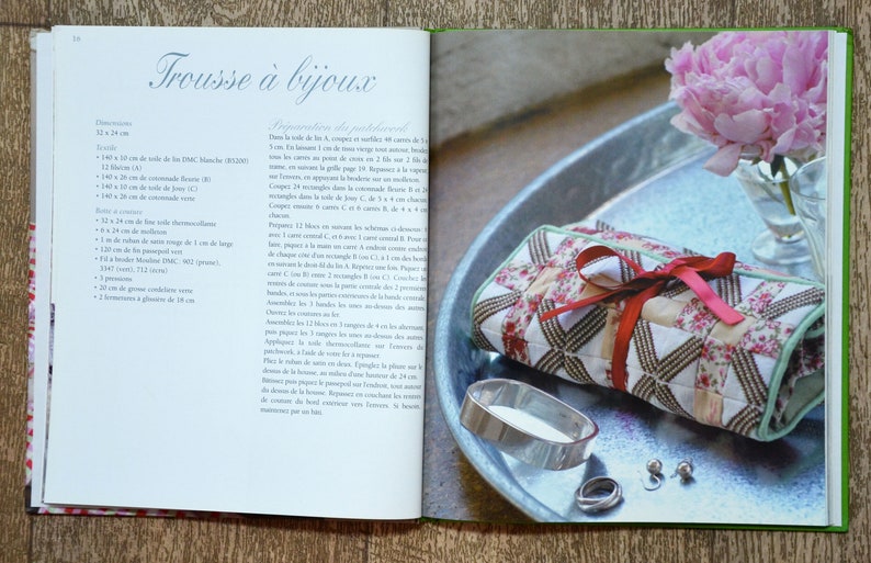 May include: A red and white quilted fabric jewellery roll with a red ribbon tied around it. The roll is sitting on a silver tray with a silver ring and two earrings. The tray is on a wooden surface with a pink flower in the background.