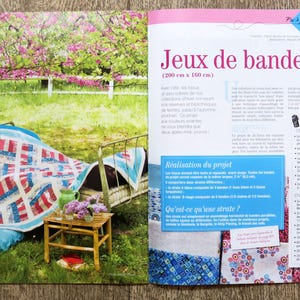 May include: A quilt made of red, white, and blue fabric strips is draped over a metal bed frame. The quilt is made up of squares and rectangles, and the fabric is arranged in a geometric pattern. The quilt is sitting in a garden setting with a wooden table and a vase of flowers.