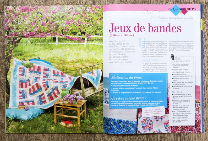 May include: A quilt made of red, white, and blue fabric strips is draped over a metal bed frame. The quilt is made up of squares and rectangles, and the fabric is arranged in a geometric pattern. The quilt is sitting in a garden setting with a wooden table and a vase of flowers.