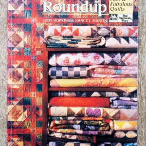 Op de afbeelding: De omslag van een boek getiteld "Rotary Roundup" met een stapel opgevouwen quilts op een houten plank. De omslag is rood met de titel in wit en de ondertitel "40 More Fast & Fabulous Quilts".
