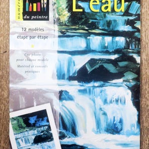 May include: A book cover with the title "L'eau" in yellow letters. The cover features a painting of a waterfall with blue and white water cascading over rocks. The book is part of a series called "Les cahiers du peintre" and includes 12 painting models.