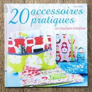 Può includere: Copertina di un libro intitolata "20 accessoires pratiques en couture créative" di Saskia Abel. La copertina presenta vari progetti di cucito, tra cui una pochette a fantasia rossa e bianca, un quaderno con un disegno di bambola e una bambola matrioska blu.