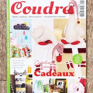Puede incluir: Una portada de revista "Coudre" que muestra proyectos de costura. La portada presenta figuras de gansos de peluche con pies y sombreros rojos, junto con otros artículos hechos a mano como cojines y un cojín en forma de rosquilla.