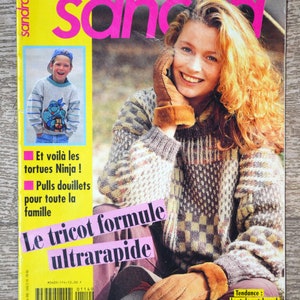 May include: Magazine cover featuring a woman with red hair wearing a patterned sweater and brown gloves. The magazine title is "sandra" with text in French. A smaller image shows a child in a sweater with a cartoon character. Text includes "Les plus beaux tricots" and "Le tricot formule ultrarapide."