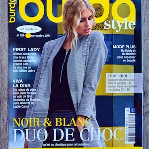 Può includere: Una copertina di rivista con una donna che indossa una giacca grigia e pantaloni neri. La rivista si chiama "burda style" e la copertina presenta il testo "Noir & Blanc Duo de Choc".