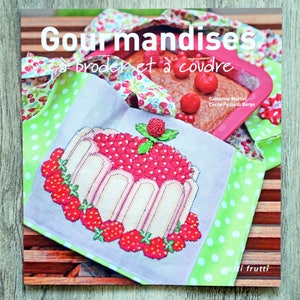Peut inclure: Un livre intitulé "Gourmandises à broder et à coudre" avec un fond en tissu à pois verts et blancs. Le livre présente un motif de point de croix d'un dessert aux fraises avec un schéma de couleurs rouge et blanc.