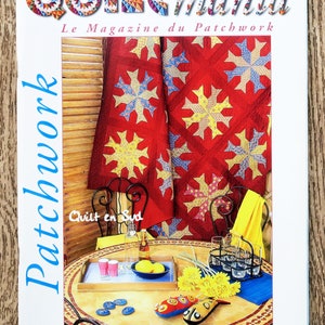 Op de afbeelding: De cover van het tijdschrift "Quiltmania" toont een rode quilt met gele en blauwe sterpatronen. De titel van het tijdschrift staat in kleurrijke letters. Het woord "Patchwork" staat verticaal aan de linkerkant van de cover.