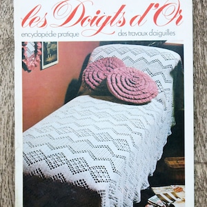 Può includere: Un copriletto bianco all'uncinetto con un motivo a rombi e bordi arricciati. Il copriletto è su un letto con due cuscini rosa all'uncinetto. La copertina della rivista recita "les Doigts d'Or encyclopédie pratique des travaux d'aiguilles".