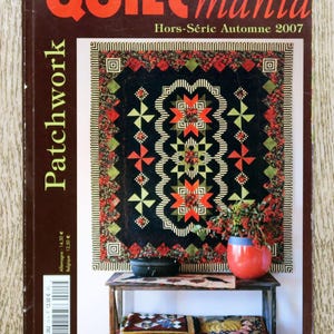 Op de afbeelding: Een quiltmagazinecover met een patchworkdeken in zwart, rood en groen. De deken hangt aan een muur boven een houten tafel met een rode vaas en andere decoratieve items. De titel van het magazine is "Quilt Mania" en de ondertitel is "Hors-Série Automne 2007".