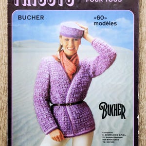 Peut inclure: Couverture de magazine vintage avec une femme portant une veste, un chapeau et une écharpe en tricot violets. Le titre est « TRICOTS FACILES POUR TOUS » et « BUCHER ». La femme sourit, avec un paysage bleu clair et blanc en arrière-plan.