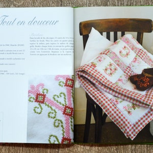 May include: A close-up of a book open to a page with the title "Tout en douceur" and a picture of a white and red gingham blanket with embroidered flowers on a wooden chair. The book is about embroidery and the blanket is a finished project.