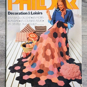 Peut inclure: Une couverture de magazine mettant en scène une femme crochetant une couverture colorée à motifs hexagonaux. Le magazine est intitulé "Phildar Décoration & Loisirs" et présente le texte "Le Ba Ba du Crochet, Les Motifs Irlandais, Les Coussins-Fleurs, La Dentelle aux Aiguilles..." en français.