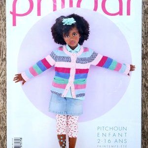 Könnte beinhalten: Ein Magazincover mit einem jungen Mädchen, das eine gestreifte rosa, blaue und grüne Strickjacke, einen Jeansrock und rote und blaue gemusterte Strumpfhosen trägt. Das Magazin heißt "Phildar" und der Text "Pitchoun Enfant 2-16 Ans Printemps-Été" ist auf dem Cover zu sehen.