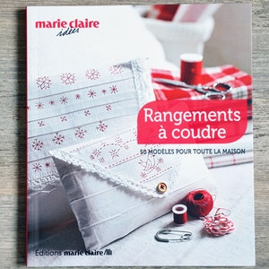 Peut inclure: Une couverture de livre avec un fond blanc et du texte rouge. Le titre est "Rangements à coudre" et le sous-titre est "50 modèles pour toute la maison". La couverture présente un oreiller blanc avec des broderies rouges et une enveloppe blanche avec des broderies rouges.